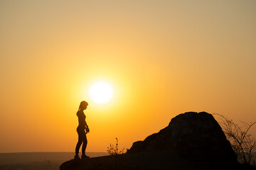 Wall Mural - Silhouette of a woman hiker standing alone near big stone at sunset in mountains. Female tourist on high rock in evening nature. Tourism, traveling and healthy lifestyle concept.