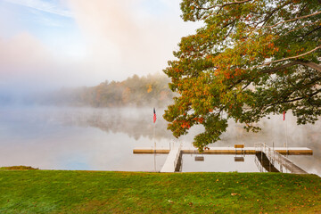 Canvas Print - Scenic river edge and T-shaped pier under oak tree early morning fog just above the water level of Connecticut River with colorful fall foliage along both sides converging into distance.