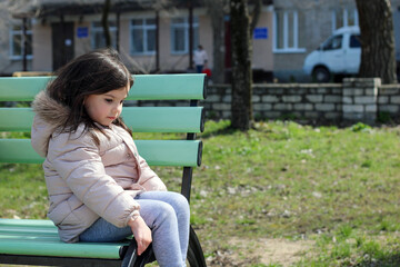 Wall Mural - Little white cute smiling girl sits on a bench against the background of a blurry building cross-legged