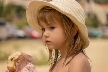 Beautiful little girl with hat eating a doughnut on the beach.