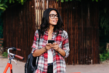 Wall Mural - Ecstatic curly girl in red checkered shirt standing in front of bicycle. Outdoor portrait of good-humoured latin lady with phone looking around the street.