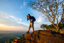 A Photographer Standing On Top Of Mountain At Sunset With Mist.Asian Male Backpack In Nature During Sunset Relax Time On Holiday Concept Travel At Pha Mor I Dang,Sisaket Province,Thailand,ASIA.