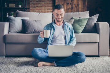 Photo of handsome homey guy relaxing sitting comfy fluffy carpet near couch drink coffee browsing notebook freelancer remote work stay home quarantine time weekend living room indoors