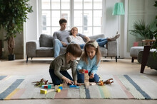 Happy Little Boy And Girl Kids Sit On Warm Floor In Living Room Building With Wooden Colorful Blocks Bricks, Young Parents Relax On Comfortable Sofa In Background, Caucasian Family Leisure Weekend