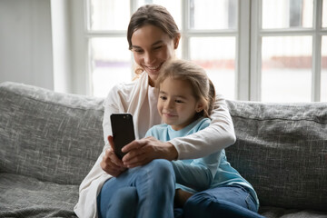 Wall Mural - Loving happy young Caucasian mom and little daughter sit relax on sofa at home smile for self-portrait picture on cellphone together, mother and small girl child make selfie on smartphone at home