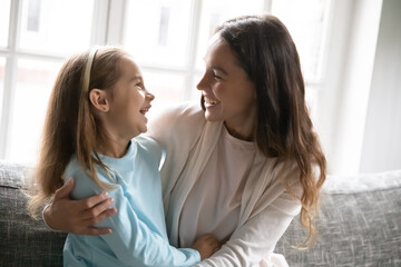 Wall Mural - Happy young Caucasian mom and little preschooler daughter sit on couch at home hugging cuddling, smiling mother or nanny rest on sofa embrace have fun play with small girl child, show love and care