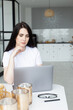 © Тетяна Шустик - Smiling young woman using laptop in the kitchen at home. Online shopping, e-learning, work from home.