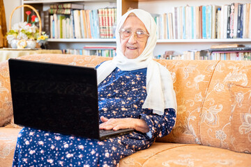 Wall Mural - Happy old arabic muslim woamn using technology while sitting on couch