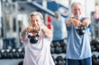 © Daniel - couple of two active and healthy seniors or pensioners or mature people doinng exercise together in the gym holding a dumbbell in their hands and doing squats - fitness lifestyle dieting concept