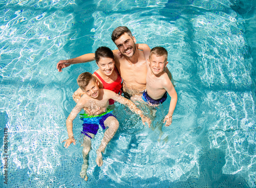 Top view of smiling family of four having fun and relaxing in indoor swimming pool at hotel resort. - obrazy, fototapety, plakaty 