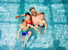 Top View Of Smiling Family Of Four Having Fun And Relaxing In Indoor Swimming Pool At Hotel Resort.