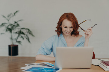 Wall Mural - Positive busy woman with European appearance works on freelance digital project, earns money online on distance job, sits at workplace with papers and notepad, wears casual shirt, holds glasses