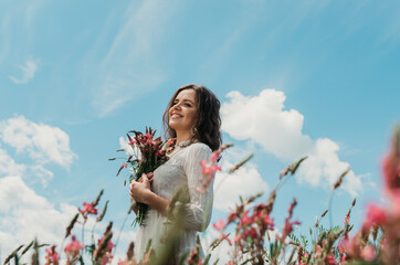 Mid body portrait of a young woman in white dress smiling to the side and holding a flower bouquet, shot against blue bright sky