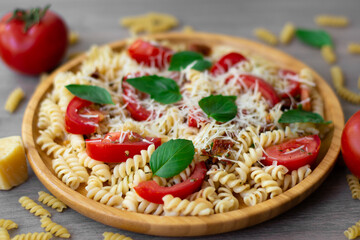 Summer cold pasta salad with fusilli pasta, fresh tomatoes, sun-dried tomatoes, grated Parmesan cheese and fresh green basil leaves in a wooden plate surrounded by tomatoes, basil, dry pasta and parm