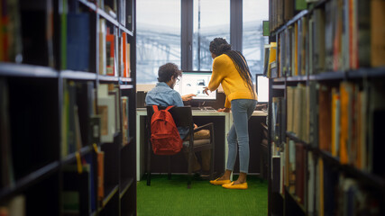 Wall Mural - University Library: Boy Uses Personal Computer at His Desk, Talks with Girl Classmate who Explains, Helps Him with Class Assignment. Focused Students Study Together. Shot Between Rows of Bookshelves