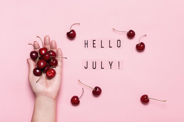 Hello July text and female hand holds ripe cherries on pink background. Hello July concept