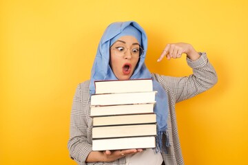 Young beautiful muslim woman wearing hijab and holding books on her arms against yellow background looking to all she has to study, being surprised and pointing at books. Education concept.