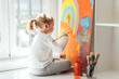 © Angel Santamaria/ADDICTIVE STOCK - Creative blond girl in casual clothes sitting on window sill against window and painting with paintbrush large multi colored rainbow on orange canvas