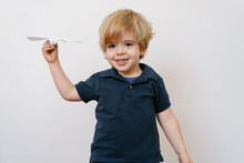Cute Blond Boy In Casual Outfit Playing With Paper Plane Smiling Brightly At Camera On White Wall Background