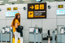 Full Length Of Joyful Female Passenger In Fashionable Outfit With Luggage And Passport In Back Pocket Looking At Camera And Waving Goodbye While Standing Next To Check In Counter At Airport
