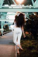 Young Slim Female In Sports Top And Leggings Standing Barefoot On Pavement Curb And Raising Arm Gracefully In Evening City