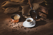 Fresh Black Coffee In White Ceramic Cup Placed On Saucer Near Coffee Grinder And Coffee Beans On Wooden Table