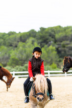 Child Looking At Camera In Jockey Costume Sitting In Saddle Of A Pony During Lesson In Horseback Riding School