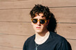© Manuel Ruiz Alba/ADDICTIVE STOCK - Modern young handsome man in trendy sunglasses and black t-shirt standing near brown wall on sunny day on city street