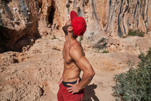 Side View Of Muscular Shirtless Bearded Climber In Red Cap Standing With Hands On Waist Near Mountain Slope And Looking Up Before Climbing
