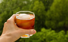 Stay At Home Balcony Cocktail Party. Classic Home-made Old Fashioned Alcohol Drink With Ice Ball And Orange Zest, Trees Sky And Rain Drops. Good For Website Blog Recipes Articles. Free Copy Space