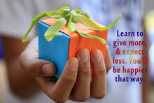 Inspirational Quote - Learn To Give More, And Expect Less. You Will Be Happier That Way. With Young Woman Hand Holding A Small Cute Gift Box With A Green Cananga Odorata Flower On It.  