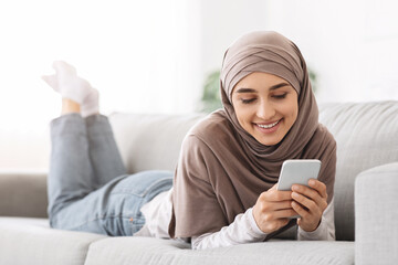 Canvas Print - Beautiful arabic girl in hijab lying on couch and texting on smartphone