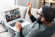 African-American young guy is using app on laptop for video meeting with many people together. Black man with a laptop lays on the couch and waving hello to group of friends on video screen