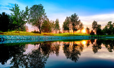 Wall Mural - summer sunset at park panorama landscape with picturesque trees and colorful sky reflection on pond water against setting sun background. Wide view of nature in russia. Mellow evening