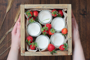 Woman holds a wooden box with 5 jars of home made yoghurt and fresh strawberries. Rustic wooden background