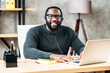 Cheerful African-American guy uses a handsfree headset and laptop to talk online at his workplace, black confident man in glasses sits at the office desk, looks at camera with pleasant smile