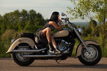 
A woman in a black polka-dot dress sits on a motorcycle against a background of trees.