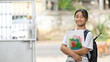 © Prathankarnpap - A schoolgirl is holding painting equipment and carrying a school bag while standing and waiting for a school bus.