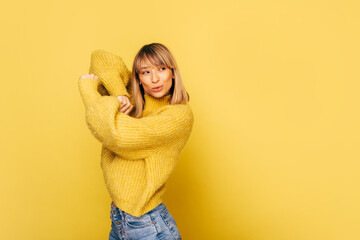 Young confident slim blonde woman hold hands up together and pose on camera. Girl stand alone in studio isolated over yellow background. Flexible fit body shape covered modern trendy jacket.