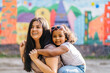 © Iryna - Mixed race laughing ecuadorian mother playing and carrying young preschooler dark-skinned daughter on her shoulders while walking together in playground. Blurred children s drawing on background.