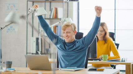 Poster - Handsome Caucasian IT Specialist Sitting at His Desk works on a Laptop, Raises Arms in Celebration after He Successfully Finished Project. Diverse Team of Young Professionals Working in Office.