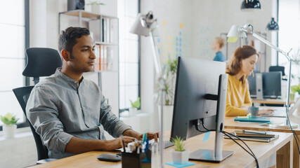 Aufkleber - Smart and Handsome Indian Office Worker Sitting at His Desk works on a Laptop. In the Background Modern Office with Diverse Team of Young Professionals Working.