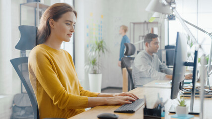 Poster - Beautiful and Creative Young Woman Sitting at Her Desk Using Laptop Computer. In the Background Bright Office where Diverse Team of Young Professionals Work.