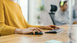 © Gorodenkoff - Anonymous Young Woman Sitting at Her Desk Using Laptop Computer. Focus on Hands Using Mouse and Keyboard. In the Background Bright Office where Diverse Team of Young Professionals Work.