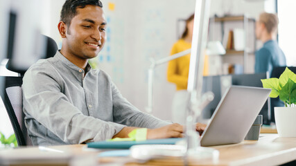 Handsome Smiling Indian Office Worker Sitting at His Desk works on a Laptop. In the Background Modern Office with Diverse Team of Young Professionals Working.
