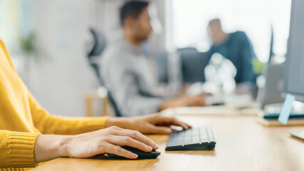 Aufkleber - Anonymous Young Woman Sitting at Her Desk Using Laptop Computer. Focus on Hands Typing on Keyboard. In the Background Bright Office where Diverse Team of Young Professionals Work.