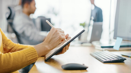 Poster - Anonymous Young Woman Sitting at Her Desk She's Drawing, Writing and Using Pen with Digital Tablet Computer. Focus on Hands with Pen. Bright Office where Diverse Team of Young Professionals Work.