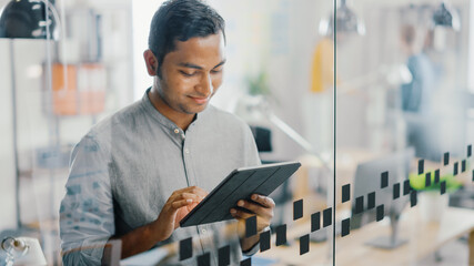 Poster - Portrait of Handsome Professional Indian Man Uses Touch Screen Digital Tablet Computer, Writes Important Email, Smiles Charmingly. Successful Man Working in Bright Diverse Office.