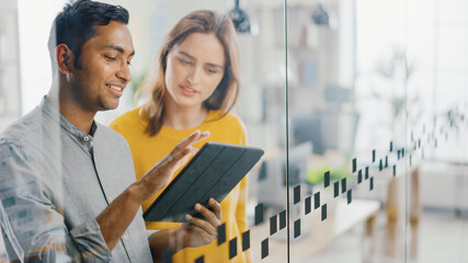 Portrait of Handsome Indian Man Showing Touch Screen Digital Tablet Computer To His Beautiful Female Team Member, They Discuss Project Details. Successful Young People Working in Diverse Office.