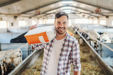 Young Smiling Attractive Caucasian Farmer Standing In Barn With Sack With Animal Food Over His Shoulder And Looking At Camera.